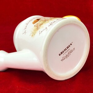 Pode incluir: Regador de cer&acirc;mica branca com al&ccedil;a amarela e design floral. O texto "A kindness is long after it is" est&aacute; impresso no lado. A parte inferior do regador tem o texto "Gretchen Stoneware" e outros detalhes.