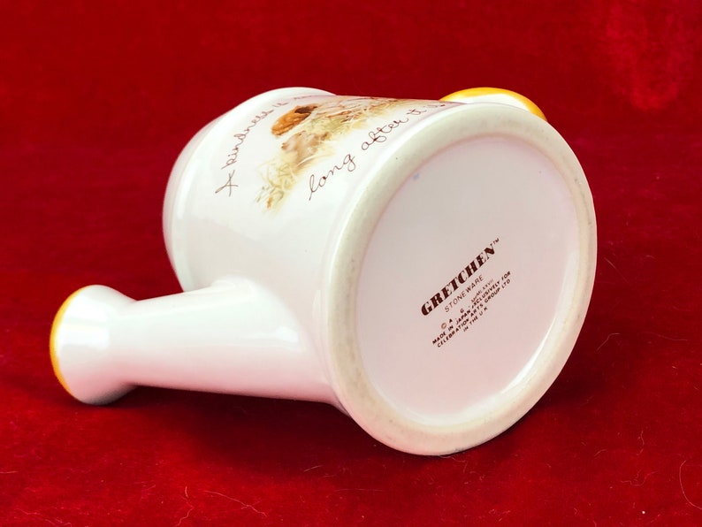 Pode incluir: Regador de cer&acirc;mica branca com al&ccedil;a amarela e design floral. O texto "A kindness is long after it is" est&aacute; impresso no lado. A parte inferior do regador tem o texto "Gretchen Stoneware" e outros detalhes.