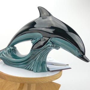Peut inclure: Une sculpture de dauphin en céramique noire brillante et turquoise, sautant au-dessus d'une vague turquoise. La sculpture est sur une petite table ronde avec une base en bois. Un bloc-notes et un stylo sont visibles.