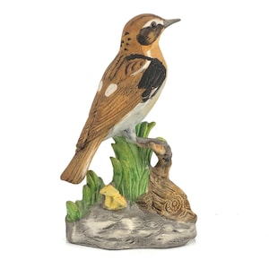 Puede incluir: Figura de pájaro de cerámica con marcas marrones, negras y blancas, posada sobre una base verde y marrón. El pájaro tiene un diseño detallado y está sobre un fondo blanco. La base incluye hierba verde y un pequeño objeto amarillo.