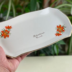 Puede incluir: Bandeja de servir de cerámica blanca con borde dorado y diseño floral. La bandeja presenta dos lirios naranjas y el texto "Saskatchewan Canada".