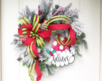 May include: A Christmas wreath featuring a red and green ribbon bow, a Santa hat with white polka dots, and the word "Believe" written on it. The wreath is adorned with frosted pine branches and red berries.