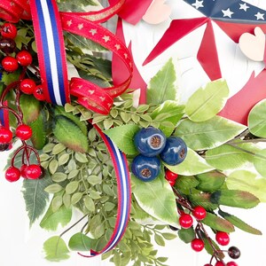 May include: A patriotic wreath featuring red, white and blue ribbon, red berries, green leaves and blue berries. The wreath includes a metal cutout of a star with red and blue accents.