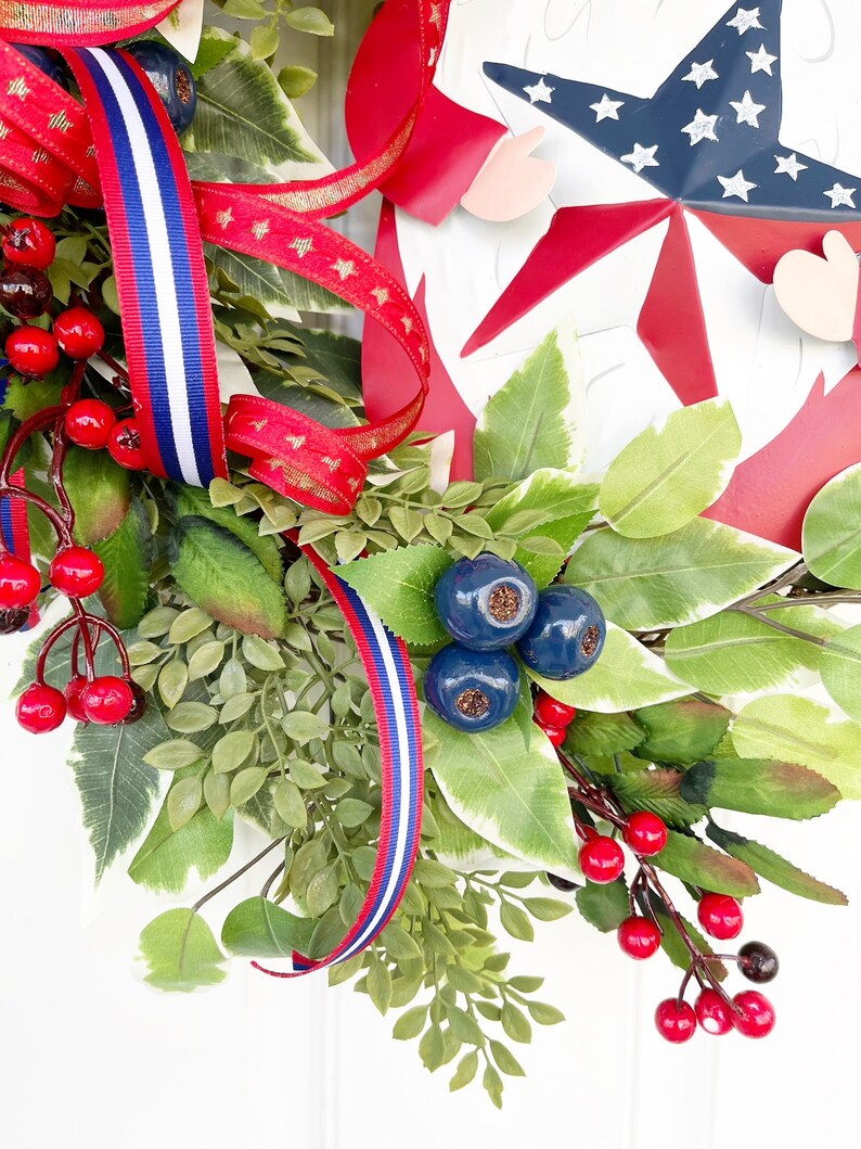 May include: A patriotic wreath featuring red, white and blue ribbon, red berries, green leaves and blue berries. The wreath includes a metal cutout of a star with red and blue accents.