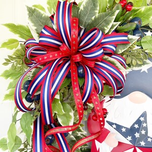 May include: A red, white and blue striped ribbon bow with gold stars is tied around a wreath of green leaves and red berries.
