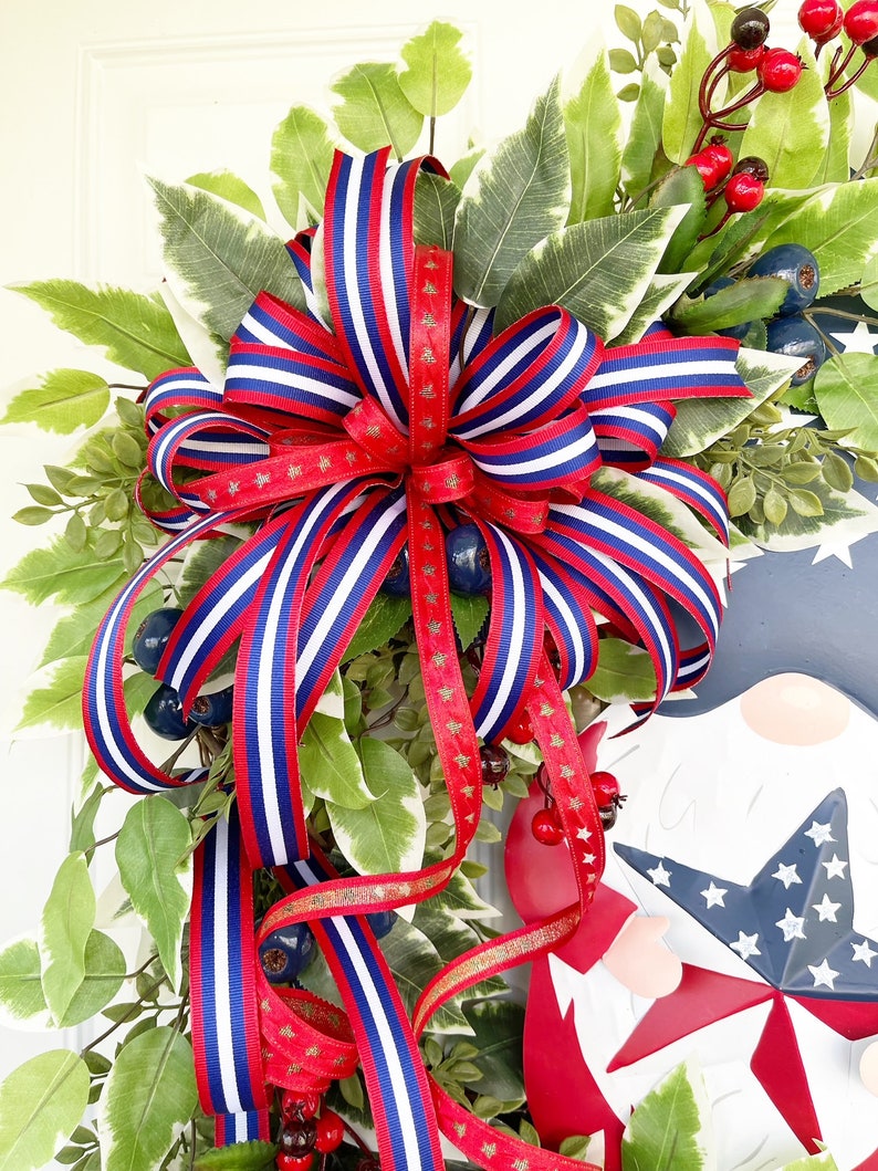 May include: A red, white and blue striped ribbon bow with gold stars is tied around a wreath of green leaves and red berries.