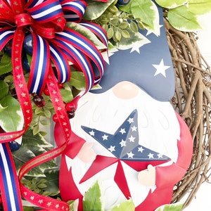 May include: A red, white and blue gnome holding a star-shaped decoration. The gnome is surrounded by greenery and a red, white and blue ribbon bow.