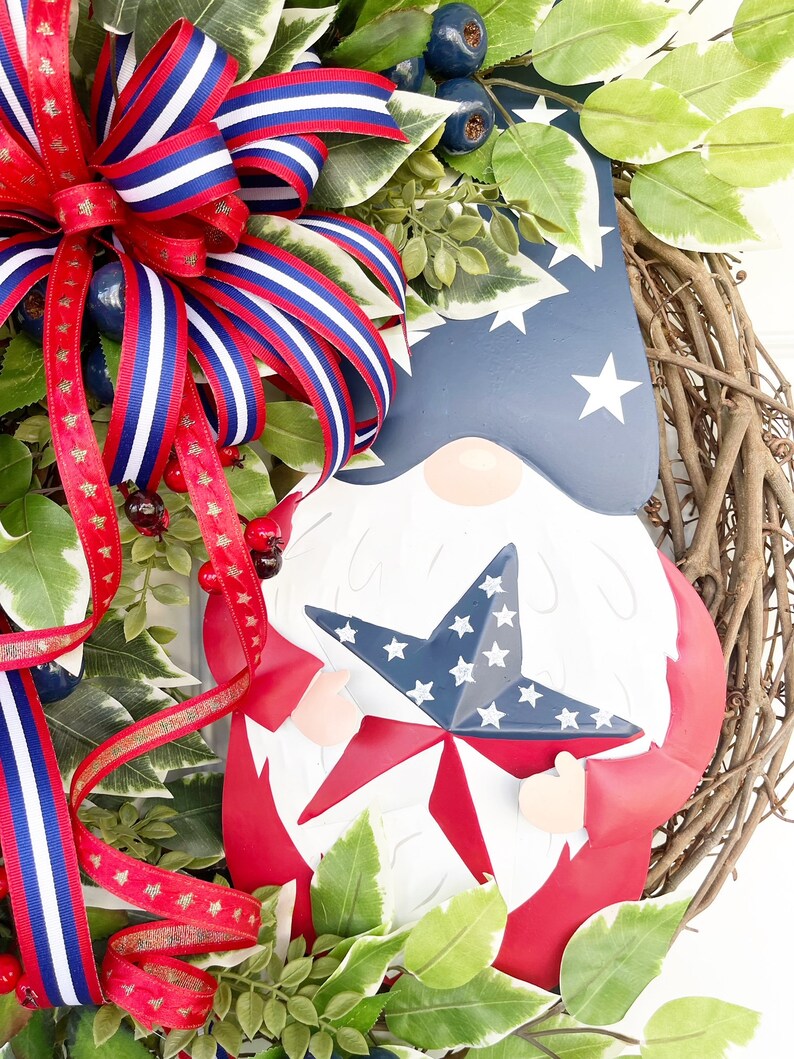 May include: A red, white and blue gnome holding a star-shaped decoration. The gnome is surrounded by greenery and a red, white and blue ribbon bow.