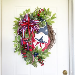 May include: A patriotic wreath featuring a gnome holding a star. The wreath is decorated with red, white and blue ribbon, green leaves and red berries.