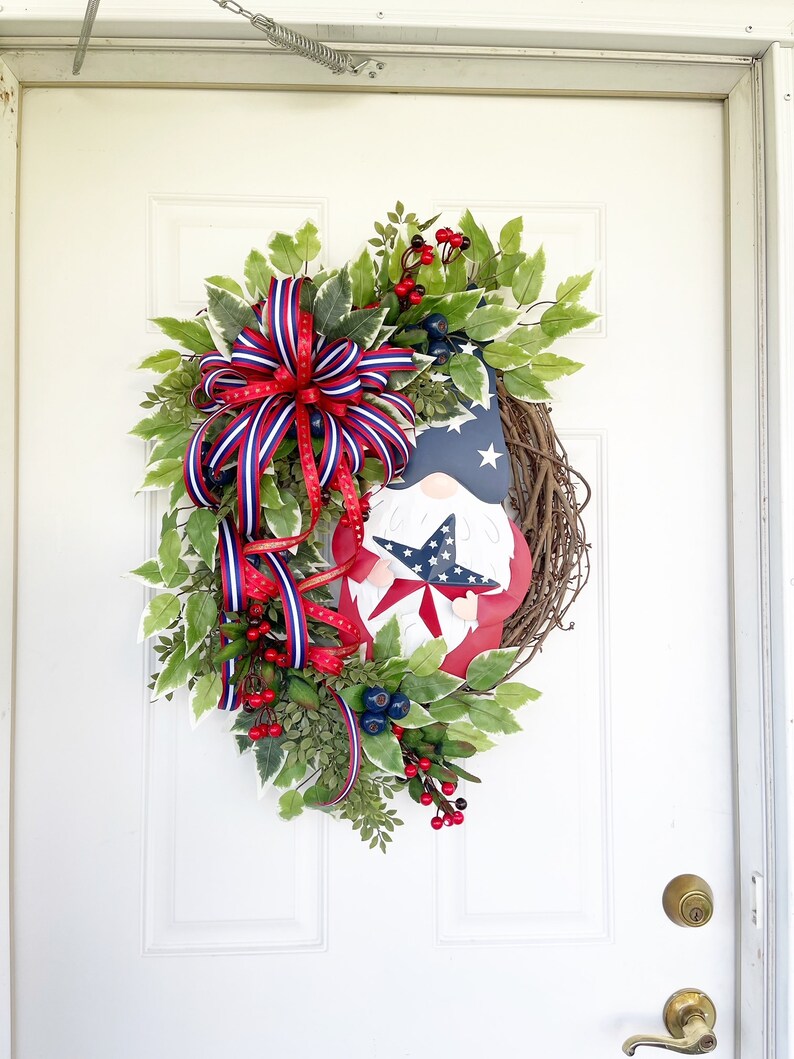May include: A patriotic wreath featuring a gnome holding a star. The wreath is decorated with red, white and blue ribbon, green leaves and red berries.