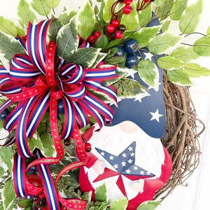 May include: A patriotic wreath featuring a red, white and blue gnome holding a star. The wreath is decorated with red berries, blue berries and green leaves. A red, white and blue ribbon is tied around the wreath.