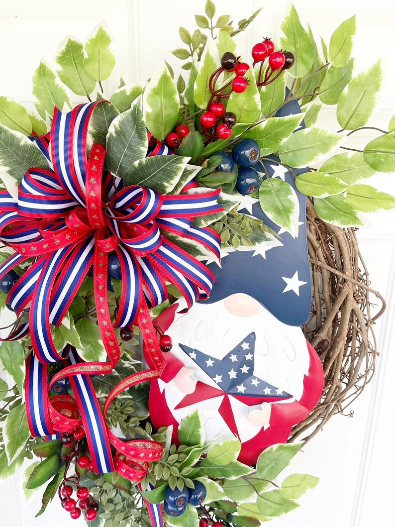 May include: A patriotic wreath featuring a red, white and blue gnome holding a star. The wreath is decorated with red berries, blue berries and green leaves. A red, white and blue ribbon is tied around the wreath.