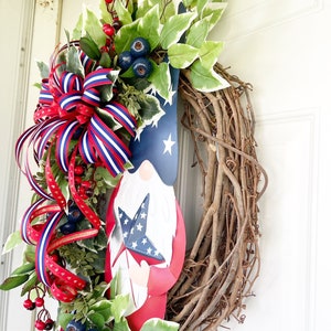 May include: A patriotic wreath featuring a red, white and blue gnome holding a star-shaped flag. The wreath is made of grapevine and decorated with red berries, blue berries and green leaves.