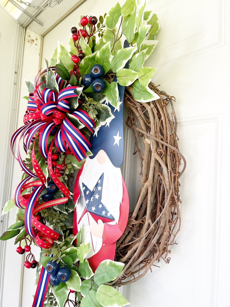 May include: A patriotic wreath featuring a red, white and blue gnome holding a star-shaped flag. The wreath is made of grapevine and decorated with red berries, blue berries and green leaves.