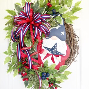 May include: A patriotic wreath featuring a gnome holding a star-shaped American flag. The wreath is decorated with red, white and blue ribbon, green leaves and red berries.