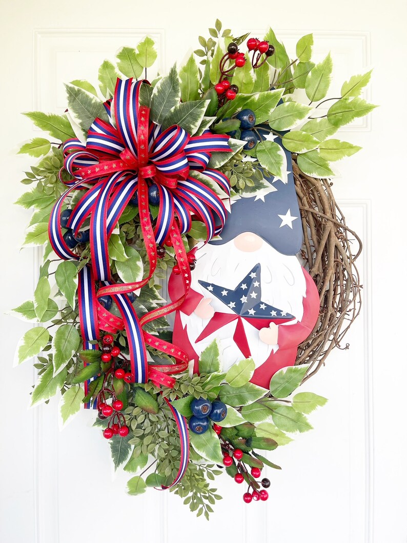 May include: A patriotic wreath featuring a gnome holding a star-shaped American flag. The wreath is decorated with red, white and blue ribbon, green leaves and red berries.