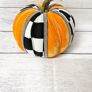 Pu&ograve; includere: Zucca di velluto arancione con un designo in tessuto a scacchi bianco e nero e strass. La zucca ha uno stelo marrone.