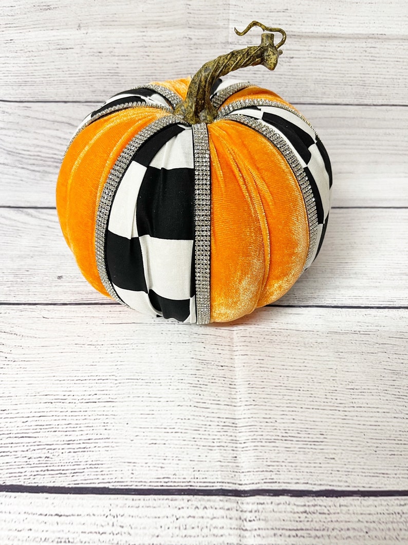 Pu&ograve; includere: Zucca di velluto arancione con un designo in tessuto a scacchi bianco e nero e strass. La zucca ha uno stelo marrone.