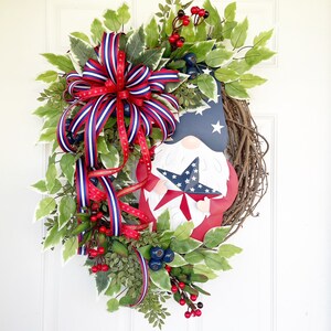 May include: A patriotic wreath featuring a red, white and blue gnome holding a star. The wreath is decorated with greenery, red berries and a red, white and blue ribbon.