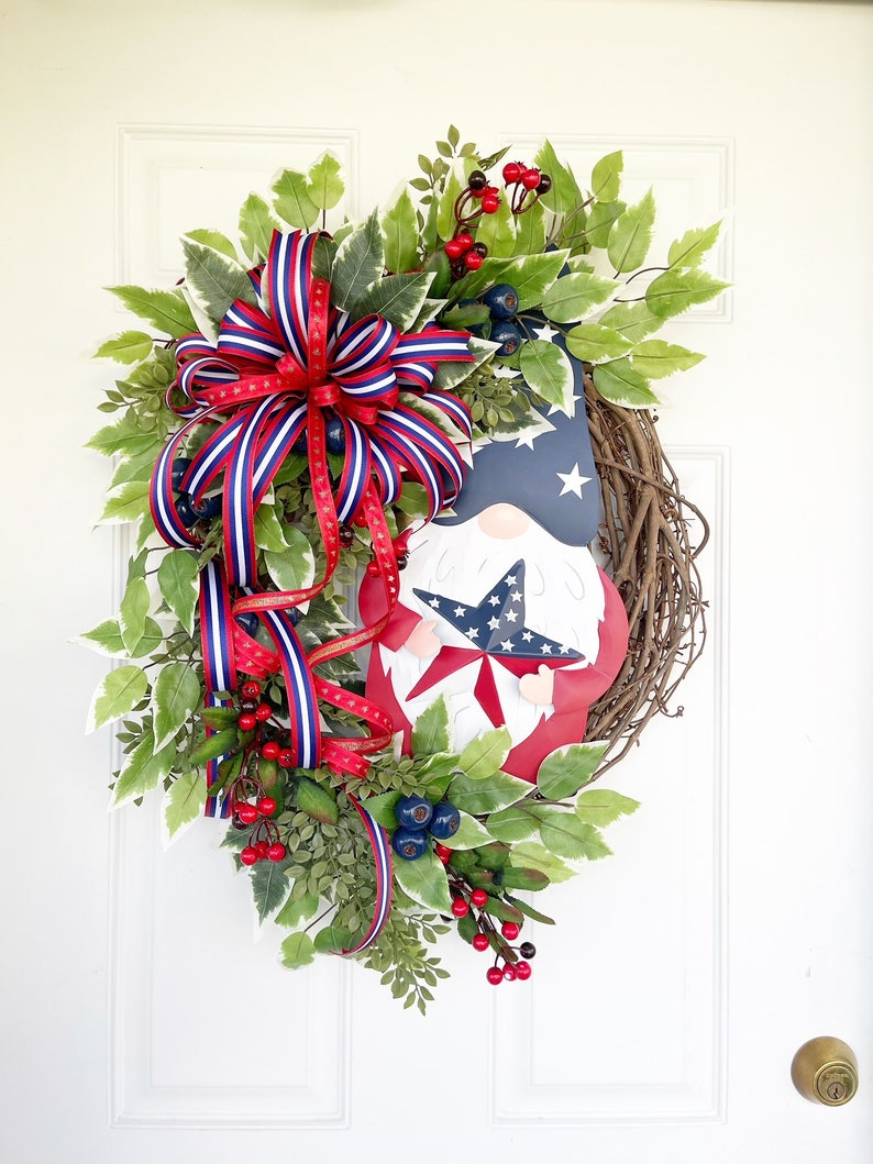 May include: A patriotic wreath featuring a red, white and blue gnome holding a star. The wreath is decorated with greenery, red berries and a red, white and blue ribbon.