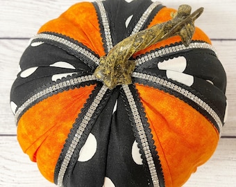 May include: A black and orange fabric pumpkin with white polka dots and a brown stem. The pumpkin is decorated with a silver ribbon with rhinestones.