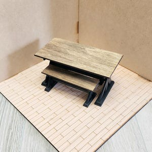 May include: A miniature rectangular table with a light brown wood grain top and a black base, accompanied by a matching smaller bench positioned directly in front of the table, both featuring a clean, simple design with sturdy black supports.