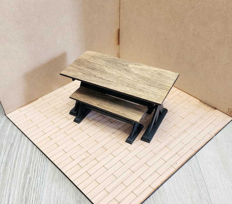 May include: A miniature rectangular table with a light brown wood grain top and a black base, accompanied by a matching smaller bench positioned directly in front of the table, both featuring a clean, simple design with sturdy black supports.