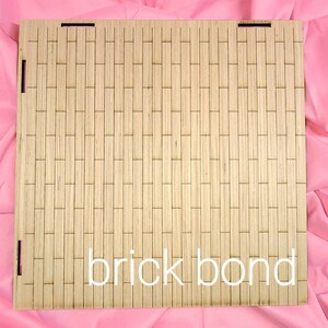 May include: A square light-colored wooden panel featuring a laser-cut pattern of vertical rectangular segments arranged in a brick bond layout. The panel includes small rectangular slots along the edges and the text brick bond in white.
