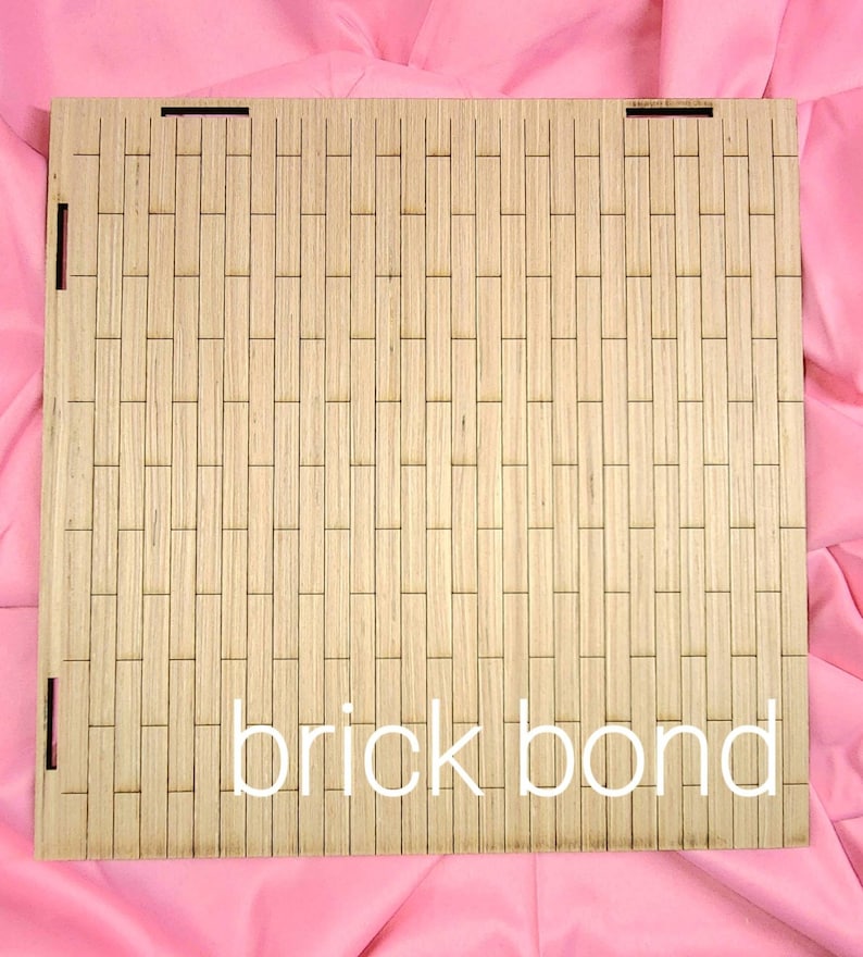 May include: A square light-colored wooden panel featuring a laser-cut pattern of vertical rectangular segments arranged in a brick bond layout. The panel includes small rectangular slots along the edges and the text brick bond in white.
