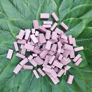 May include: A collection of small, rectangular, light purple blocks scattered on a bed of large, green leaves. The blocks appear to be made of a soft material, and are of varying sizes. The leaves provide a contrasting background, enhancing the color of the blocks.