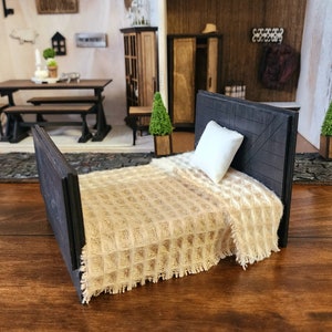 May include: A miniature black wooden bed with a tan woven blanket and a white pillow. The bed is in a dollhouse setting.