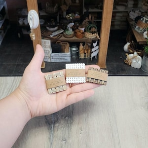 May include: Three miniature placemats in a hand. One is woven beige, one is white with square holes, and one is woven brown. Each has a brown paper band that reads "PLACEMATS SET OF 2".