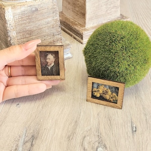 Puede incluir: Dos pequeños marcos de madera con un acabado de madera marrón. El marco de la izquierda tiene un retrato de un hombre con barba. El marco de la derecha tiene una pintura de bodegón de fruta amarilla.