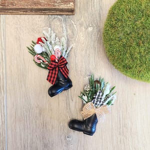 May include: Two black boot-shaped Christmas ornaments, each filled with festive greenery, candy canes, and a decorative bow. One bow is red and black plaid, the other is tan. The ornaments are on a light wood surface.