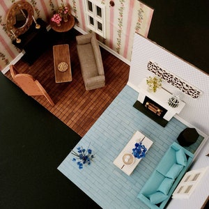 May include: A miniature dollhouse room set featuring two distinct living areas. One section has pink floral wallpaper, a brown sofa, a wooden coffee table, and a fireplace. The other section has light blue flooring, a turquoise sofa, and a white table.