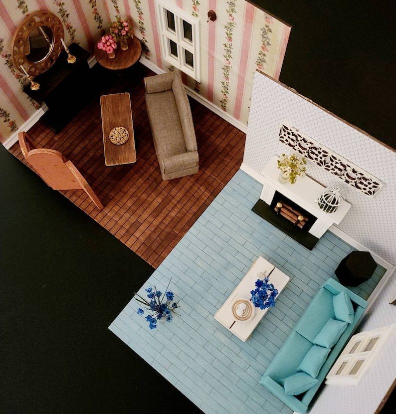 May include: A miniature dollhouse room set featuring two distinct living areas. One section has pink floral wallpaper, a brown sofa, a wooden coffee table, and a fireplace. The other section has light blue flooring, a turquoise sofa, and a white table.