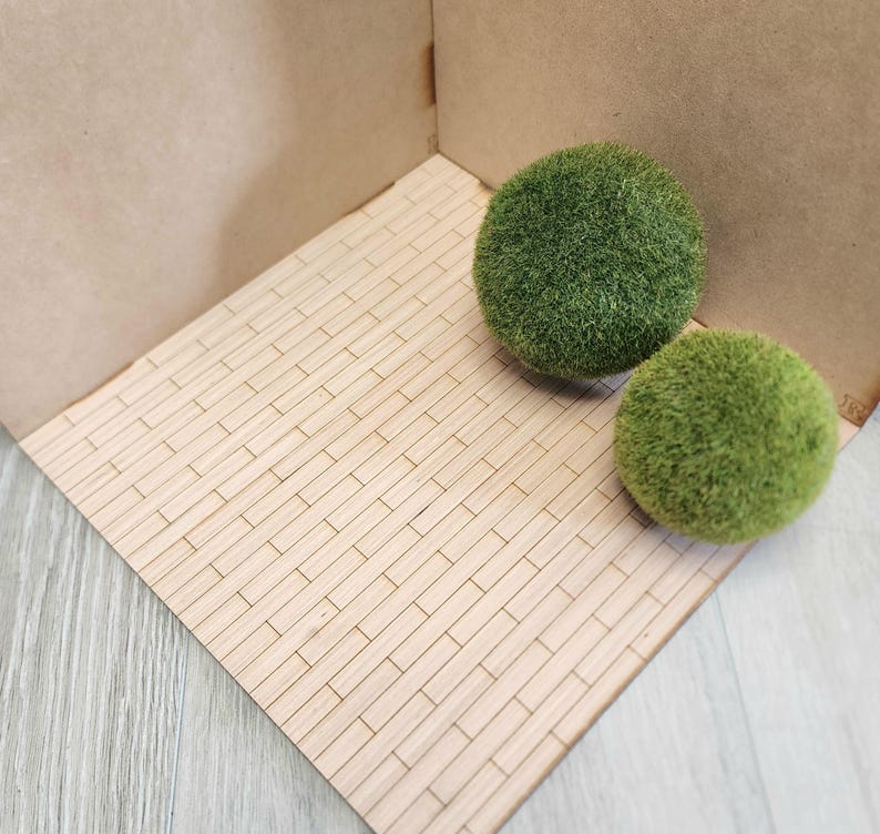 May include: Two spherical, textured green moss balls resting on a light-colored, rectangular wooden platform featuring a grid-like pattern of small, thin, light-toned wooden slats arranged in a brick-like layout against a plain backdrop.