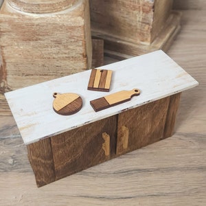 May include: A miniature wooden kitchen cabinet with two doors and gold handles. The cabinet has a white countertop with three wooden kitchen utensils: a round cutting board, a rectangular cutting board, and a small wooden spatula.