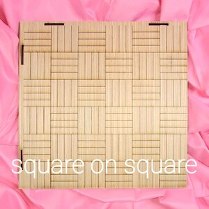 May include: A square wooden panel featuring a grid pattern of alternating vertical and horizontal slats. The light brown wood surface displays a geometric texture. The text "square on square" is printed in white at the bottom of the panel.
