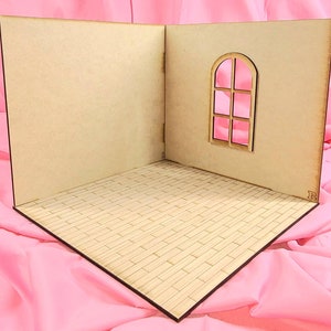 May include: A miniature room display set made of light-colored wood, featuring two perpendicular walls and a floor with a brick-patterned texture. One wall includes an arched window frame with a cross-bar design. The structure has small letters L and R.