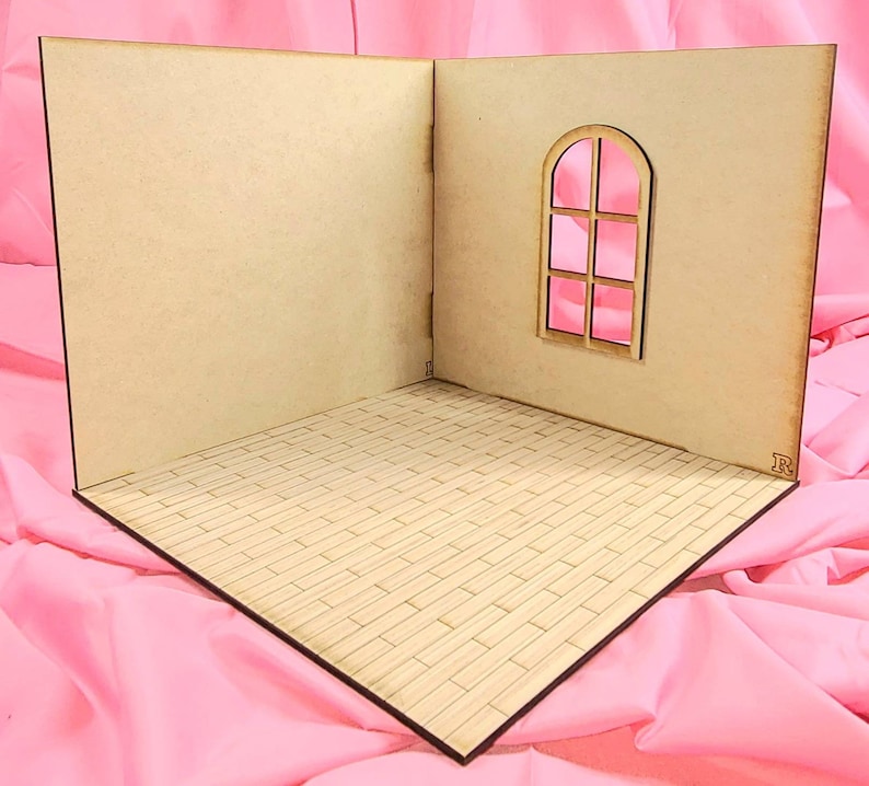 May include: A miniature room display set made of light-colored wood, featuring two perpendicular walls and a floor with a brick-patterned texture. One wall includes an arched window frame with a cross-bar design. The structure has small letters L and R.