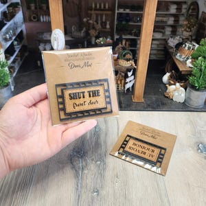 May include: Miniature door mats with black frames and cork centers. One mat reads "SHUT THE front door" and the other reads "BONJOUR AU REVOIR". The mats are packaged on brown cards with the text "Sealed Realm Miniature Crafts and Creations Door Mat".