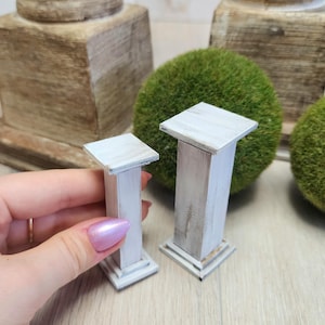 May include: Two white wooden pedestals, one taller than the other, with a distressed finish. The pedestals are square and have a simple design.
