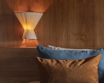 May include: A bed with a blue linen duvet cover and three pillows. The top pillow is brown, the middle pillow is blue, and the bottom pillow is white. A wall-mounted light fixture with a white shade is on the wall behind the bed.