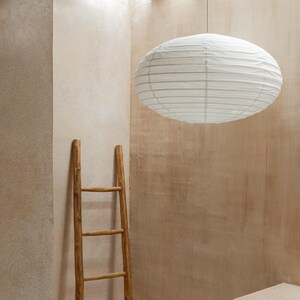 May include: A white oval paper lantern with horizontal ribbing suspended from the ceiling by a thin cord, positioned above a tall, slender wooden ladder with four rungs, featuring a natural light brown finish and a simple, rustic construction.