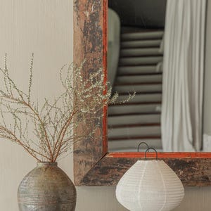 A beautifully styled boho-chic interior featuring a handcrafted linen table lamp with a soft, organic glow. The lamp sits atop two stacked design books on a rustic wooden console table, accompanied by an artisanal textured vase with delicate branches. A vintage wooden mirror with distressed edges reflects a cozy, neutral-toned space in the background. The scene exudes warmth, simplicity, and timeless craftsmanship.