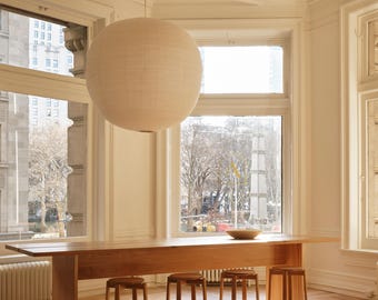 May include: A large, spherical, off-white paper pendant light hangs above a long, rectangular wooden table. Four wooden stools with circular seats and metal footrests are positioned along the table, which sits on a light-colored wooden floor.