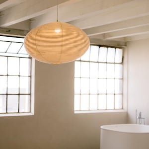 May include: A large, oval-shaped pendant lamp featuring a ribbed, off-white paper shade. The light fixture hangs from a thin cord attached to a white ceiling beam, emitting a soft, warm glow through the textured, translucent paper material.