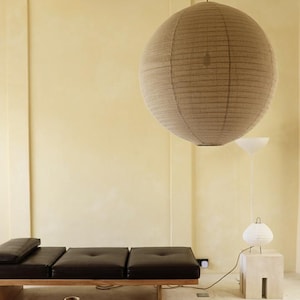 Op de afbeelding: Een modern interieur met een grote, ronde, beige papieren lantaarn die aan het plafond hangt. Daaronder een lage, donkere houten salontafel en een lange, zwarte leren bank. Een witte lamp en een kleine bijzettafel maken de scène compleet.