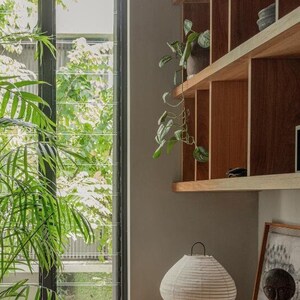The Frank Table Lamp, a handcrafted linen lantern-style lamp with a black metal frame, sits elegantly on a wooden desk in a warm, nature-inspired home office. Its soft, diffused light complements the earthy tones and Japandi aesthetic, perfect for creating a cozy and stylish workspace.